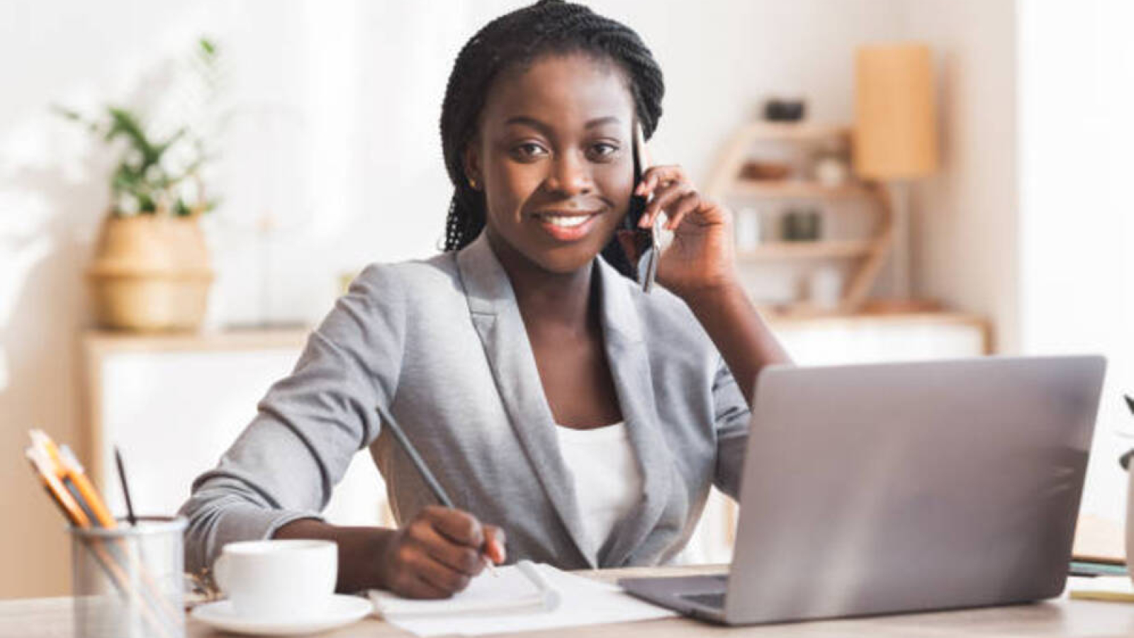 Portrait of black female entrepreneur talking on mobile phone, taking notes and smiling to camera